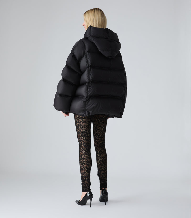 A woman viewed from behind wearing a black oversized puffer jacket, black lace leggings, and black high heels against a light gray studio background.