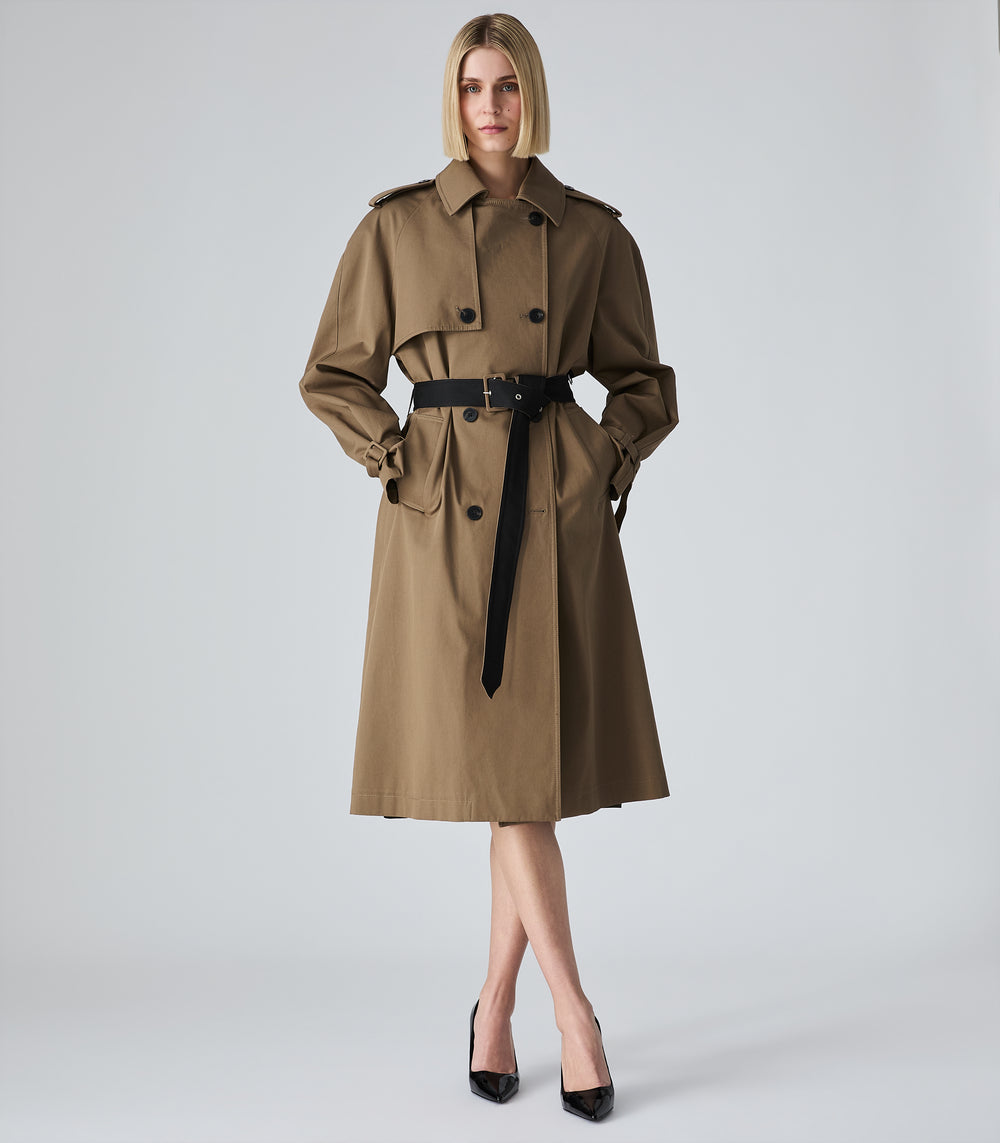 A woman modeling a classic trench coat in a warm brownish-beige color with a black belt, standing against a light gray studio background.