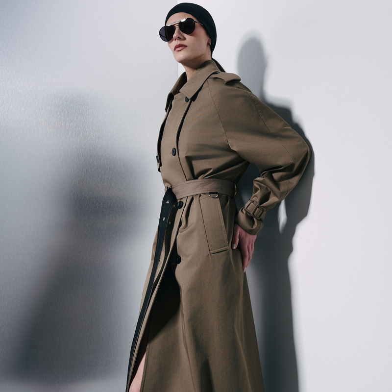 A model wearing a long olive-brown trench coat with a matching belt, black sunglasses, and a black beanie, standing against a white wall with a soft shadow.