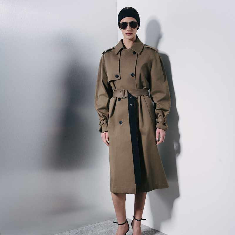 A model wearing a brown trench coat with a matching belt, black sunglasses, and a black beanie, standing against a white wall with a soft shadow.