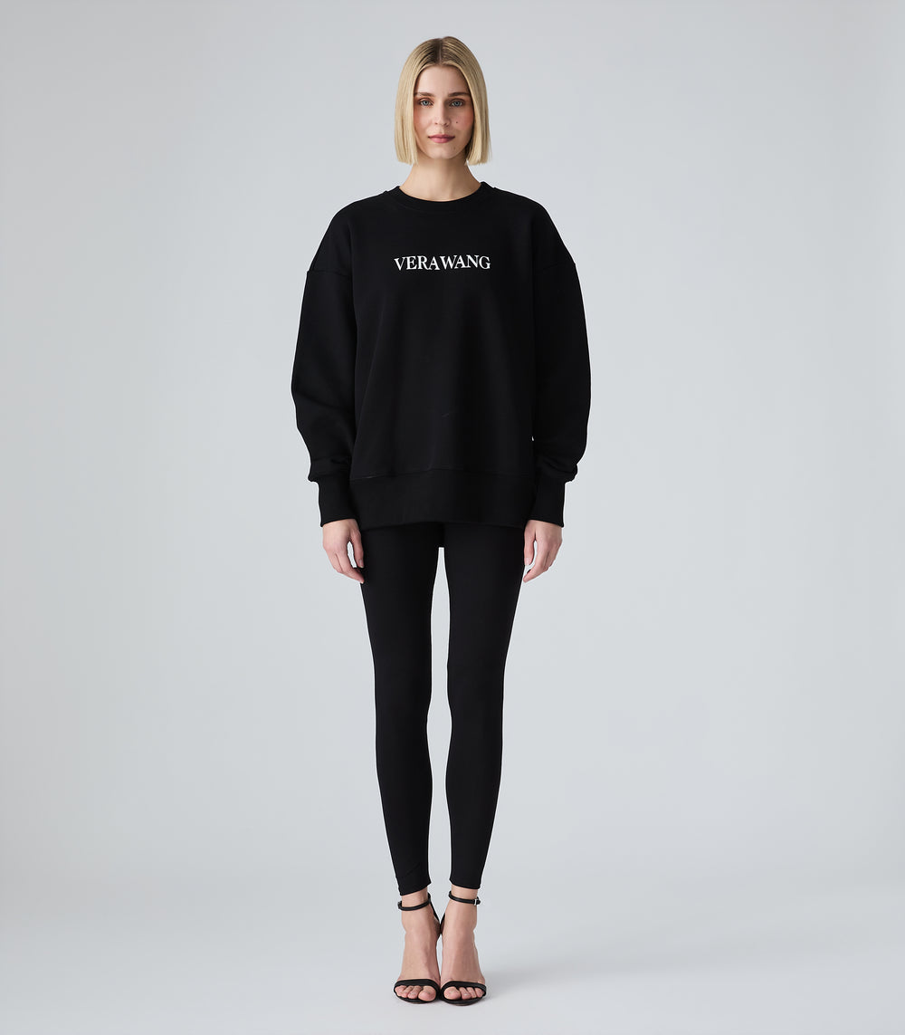 Female model wearing a black crewneck sweatshirt with 'VERA WANG' spell-out logo across the chest, paired with black leggings and heels, against a white background.