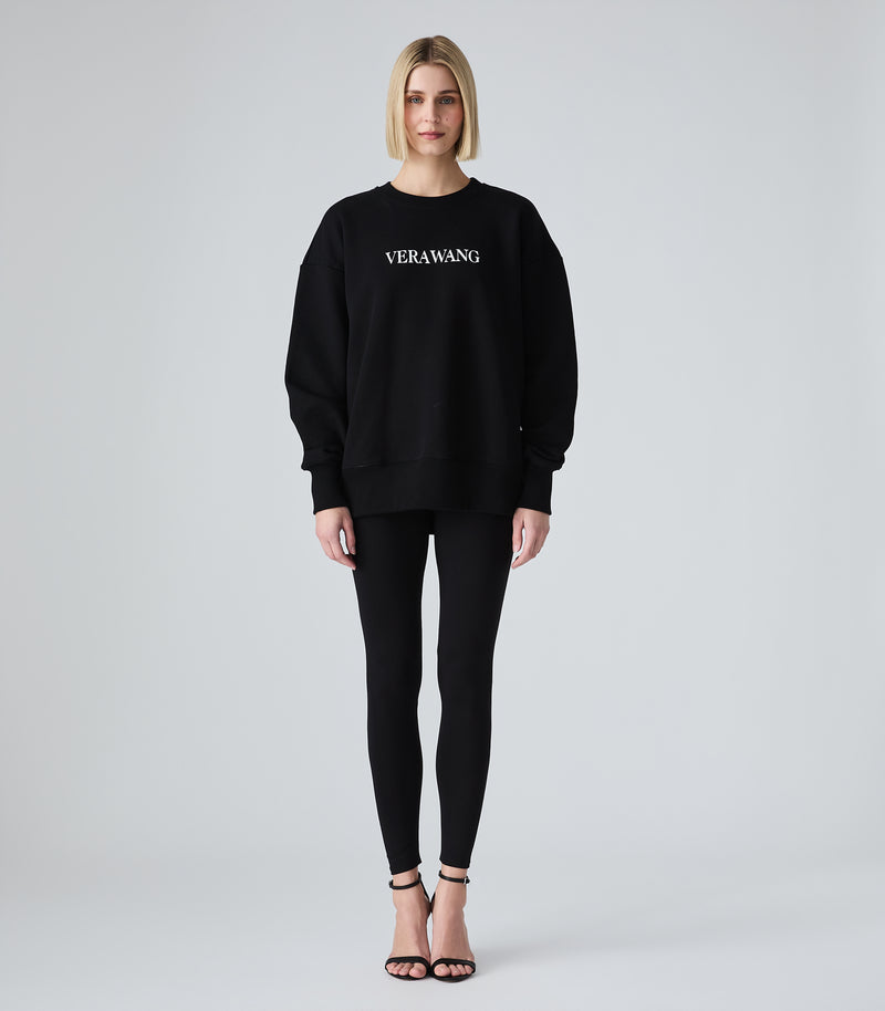 Female model wearing a black crewneck sweatshirt with 'VERA WANG' spell-out logo across the chest, paired with black leggings and heels, against a white background.