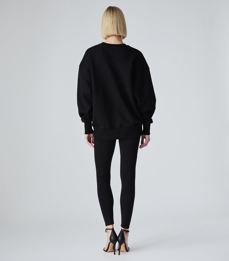 Female model wearing a black crewneck sweatshirt, paired with black leggings and heels, against a white background.