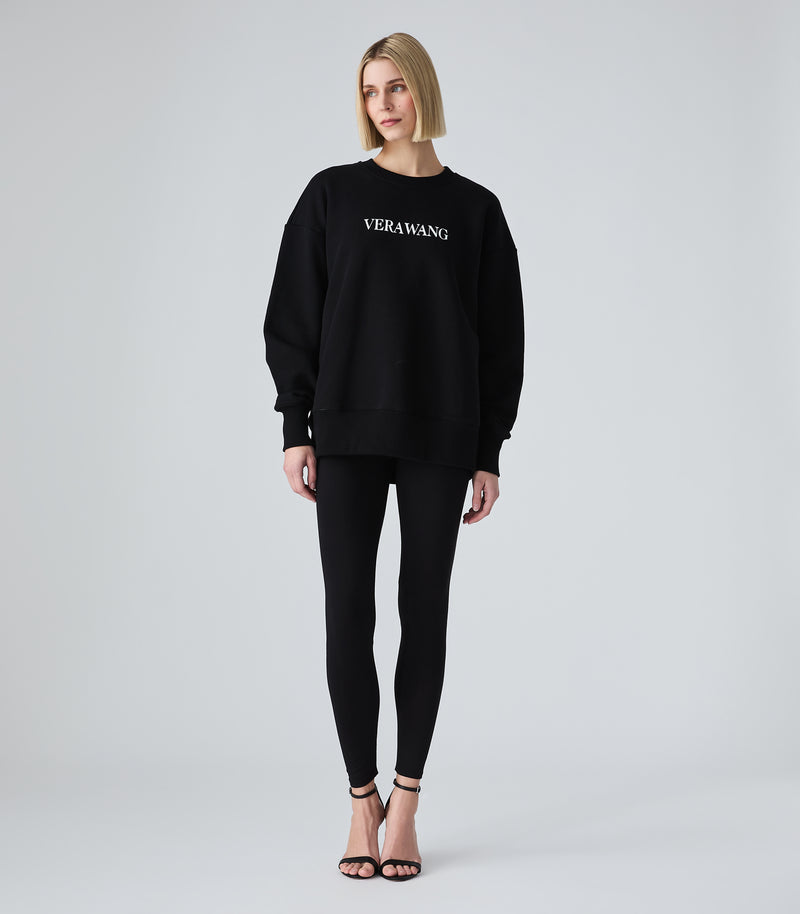 Female model wearing a black crewneck sweatshirt with 'VERA WANG' spell-out logo across the chest, paired with black leggings and heels, against a white background.