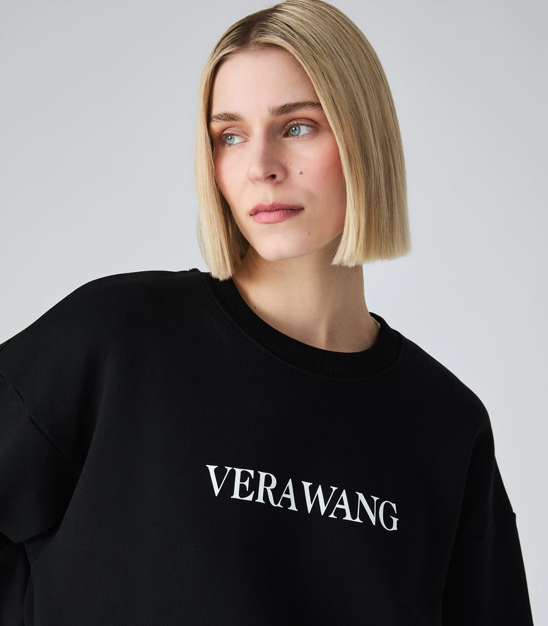 Female model wearing a black crewneck sweatshirt with 'VERA WANG' spell-out logo across the chest, against a white background.