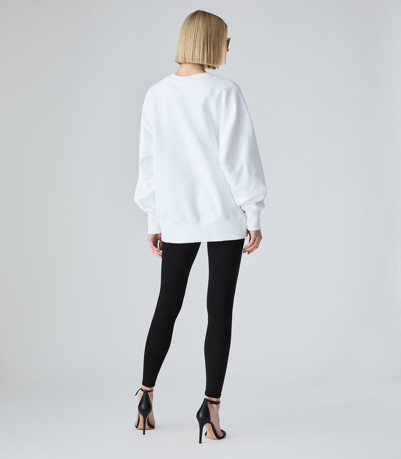 Female model wearing a white crewneck sweatshirt, paired with black leggings and heels, against a white background.