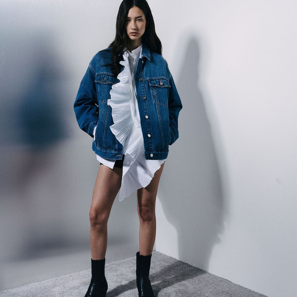 A model stands against a white wall wearing an oversized blue denim jacket over a white ruffled shirt and skirt, paired with black ankle boots.