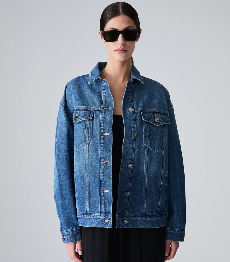 A model wearing an oversized blue denim jacket over a black top and black pants, styled with black sunglasses, against a solid light gray background.