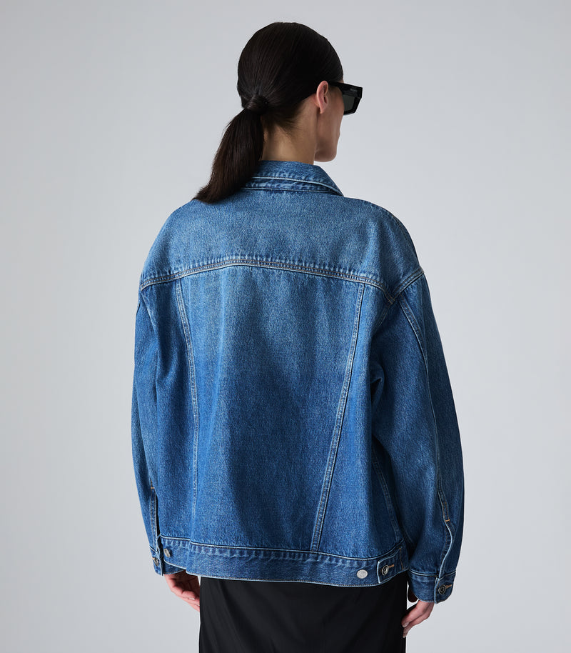 A person shown from the back wearing an oversized blue denim jacket and black pants against a light gray studio background.