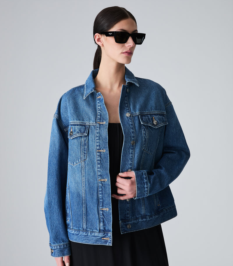 A woman modeling an oversized medium-wash blue denim jacket over a black top and black skirt, wearing black sunglasses, against a light gray studio background.