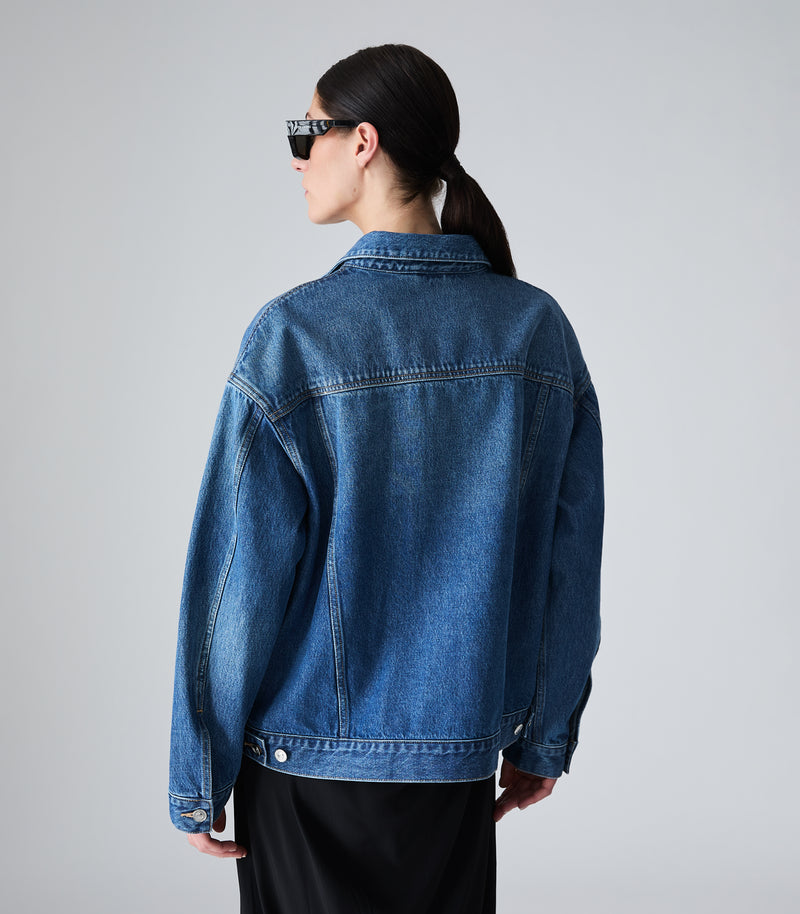 A model shown from the back wearing an oversized blue denim jacket and black pants against a light gray studio background.