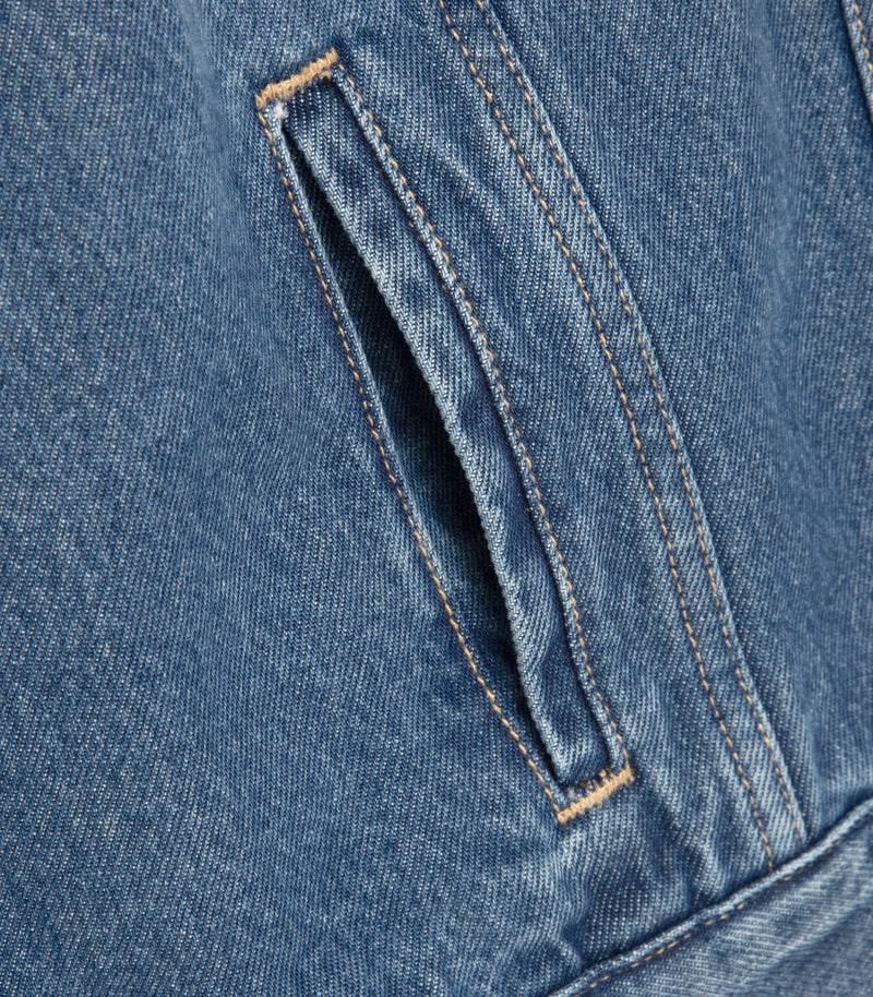 Close-up of blue denim fabric showing a diagonal welt pocket with contrast tan stitching.