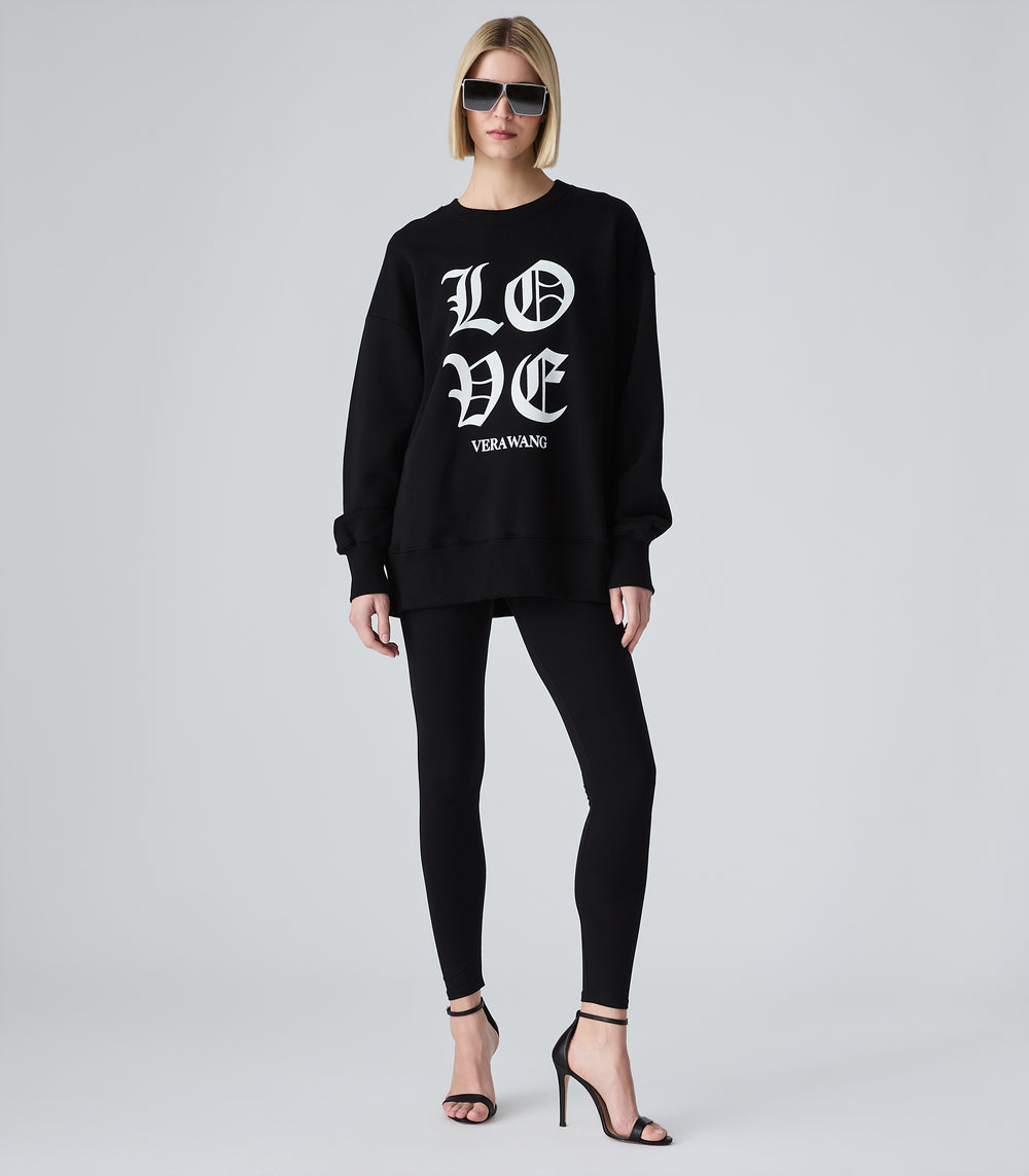 Female model wearing a black crewneck sweatshirt with 'LOVE VERA WANG' spell-out logo across the chest, paired with black leggings, sunglasses and heels, against a white background.
