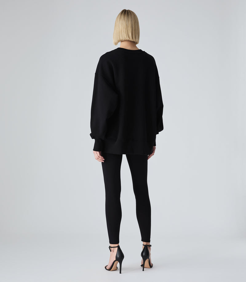 Female model wearing a black crewneck sweatshirt, paired with black leggings and heels, against a white background.