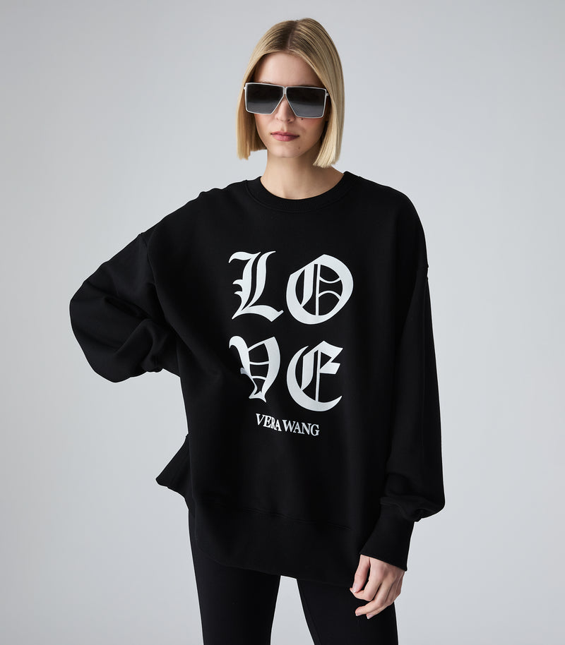 Female model wearing a black crewneck sweatshirt with 'LOVE VERA WANG' spell-out logo across the chest, paired with black leggings and sunglasses, against a white background.