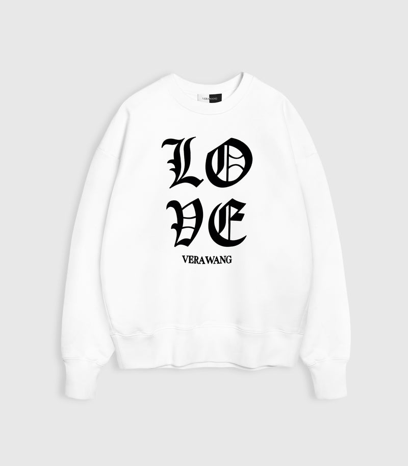 Flat lay product view of an oversized white crewneck sweatshirt featuring 'LOVE' printed in large black Old English gothic lettering with the 'VERA WANG' brand logo directly underneath, complete with ribbed cuffs and hem against a light grey background.