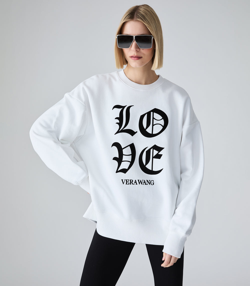 Full-length view of a model wearing an oversized white crewneck sweatshirt featuring 'LOVE' printed in large black Old English gothic lettering, with the 'VERA WANG' brand logo beneath it. The top is styled with black leggings and bold, futuristic silver shield sunglasses against a light grey background.
