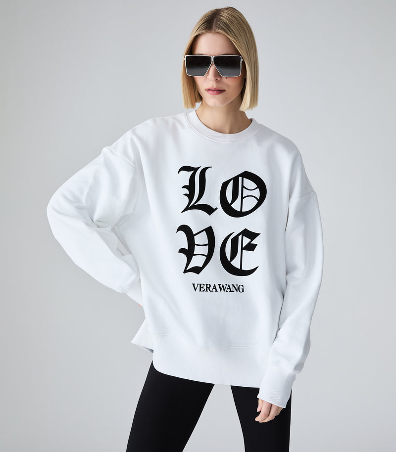 Full-length view of a model wearing an oversized white crewneck sweatshirt featuring 'LOVE' printed in large black Old English gothic lettering, with the 'VERA WANG' brand logo beneath it. The top is styled with black leggings and bold, futuristic silver shield sunglasses against a light grey background.