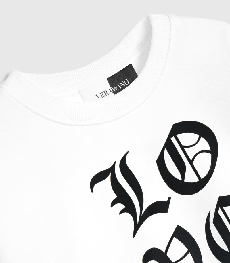 Close-up, angled product view of a white crewneck sweatshirt, showcasing the large black Old English gothic lettering of the 'LOVE' graphic across the front, a ribbed knit collar, and a woven black-and-white inner neck brand label against a light grey background.