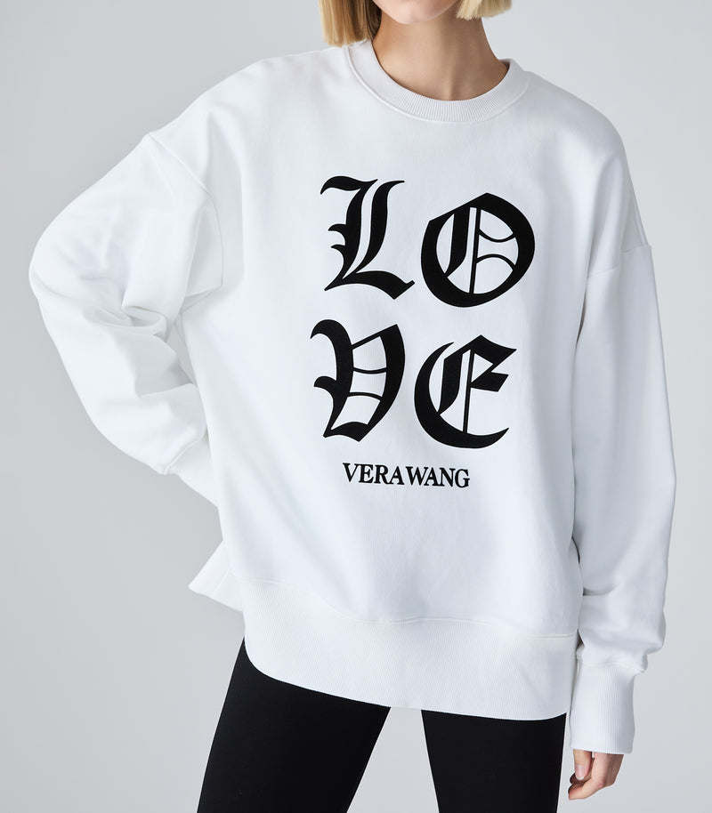 Mid-length view of a model showcasing an oversized white crewneck sweatshirt. The top features a large 'LOVE' graphic printed in black Old English gothic font with the 'VERA WANG' brand logo positioned directly underneath, styled with black leggings against a light grey background.
