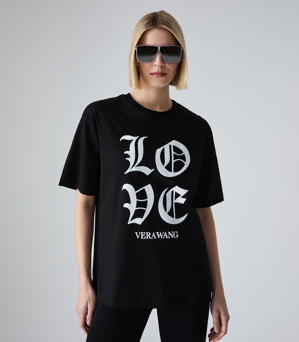 Half-length view of a model wearing a loose-fit black t-shirt featuring 'LOVE' printed in large white Old English gothic lettering with the 'VERA WANG' brand logo below it. The top is styled with black leggings, bold silver shield sunglasses, against a light grey background.