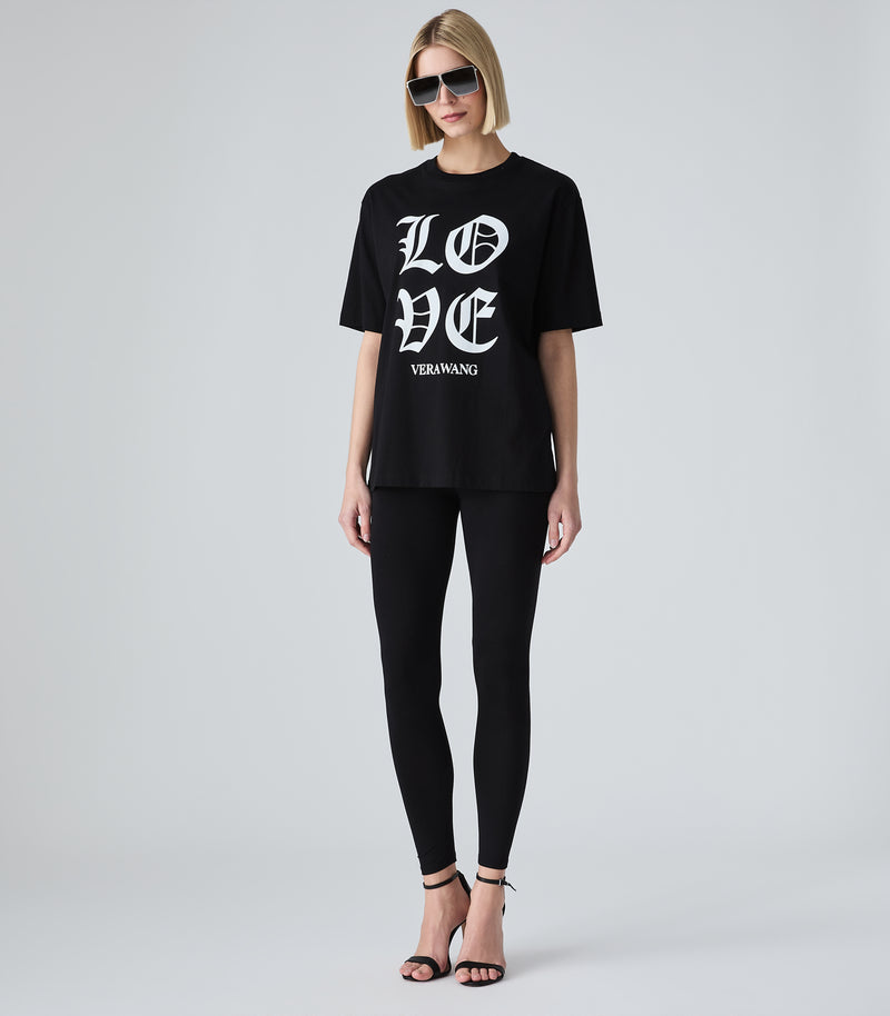 Full-length view of a model wearing a loose-fit black t-shirt featuring 'LOVE' printed in large white Old English gothic lettering with the 'VERA WANG' brand logo below it. The top is styled with black leggings, bold silver shield sunglasses, and black stiletto heeled sandals against a light grey background.