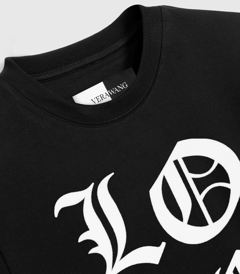 Close-up, angled product view of a black crewneck t-shirt, showcasing the large white Old English gothic lettering of the 'LOVE' graphic across the front, a finely ribbed knit collar, and a woven black-and-white inner neck brand label against a light grey background.