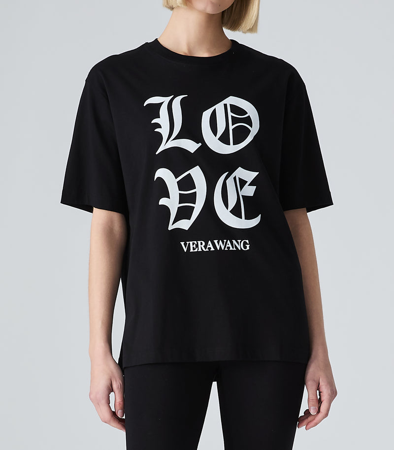 Half-length mid view of a model wearing a loose-fit black t-shirt featuring 'LOVE' printed in large white Old English gothic lettering with the 'VERA WANG' brand logo below it. The top is styled with black leggings against a light grey background.