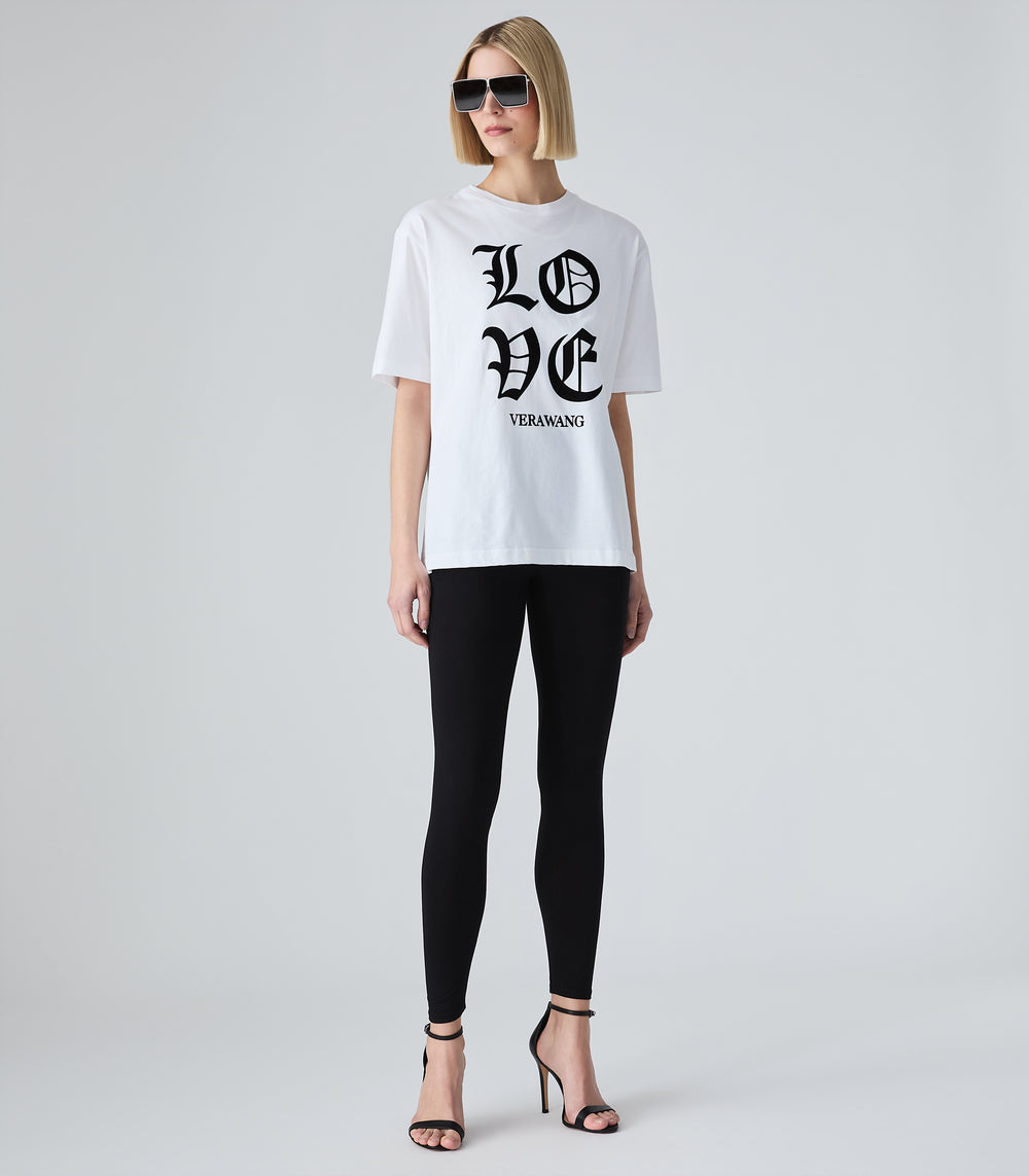 Female model wearing a white t-shirt with 'LOVE VERA WANG' spell-out logo across the front, paired with black leggings, sunglasses and heels, against a white background.