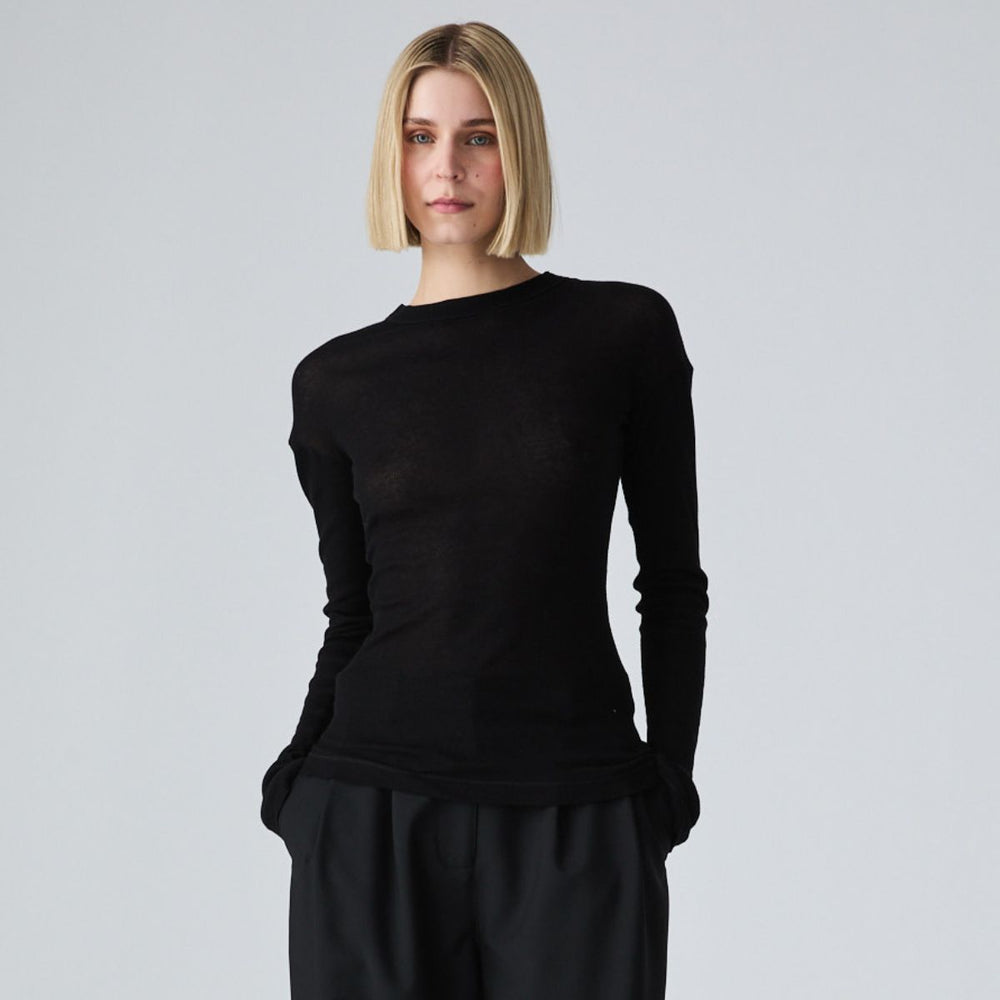 A woman with a short blonde bob wearing a black long-sleeve crewneck knit top and black trousers, standing against a light gray studio background.