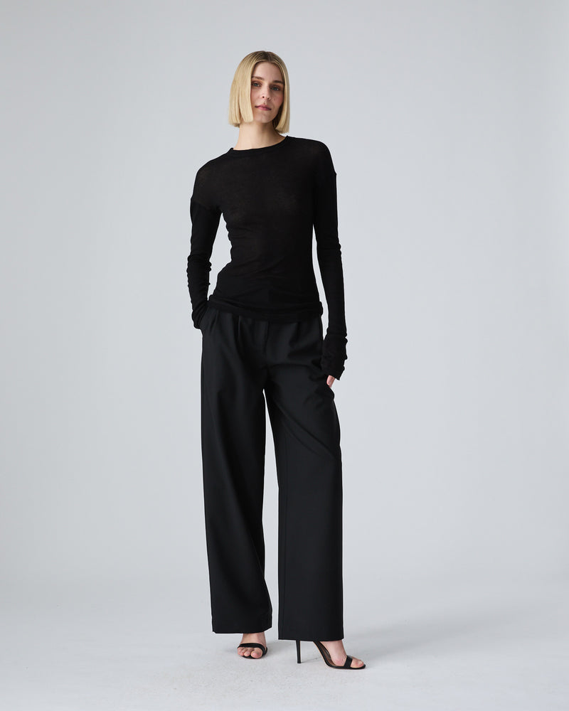 A blonde model wearing a black long-sleeve crewneck top and matching black wide-leg trousers, standing against a light gray studio background.