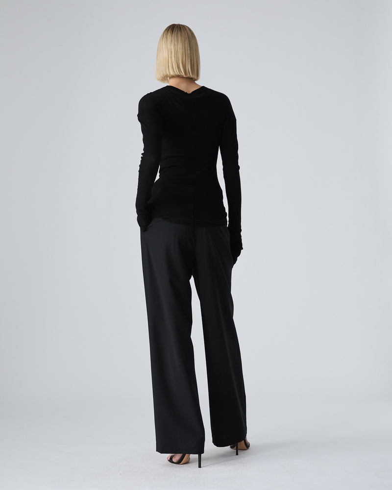A woman with a short blonde bob is shown from the back wearing a black long-sleeve top and black wide-leg trousers, standing against a light gray studio background.