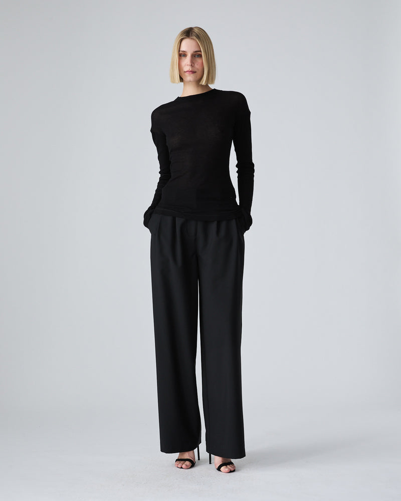 A woman modeling a black long-sleeve top and black wide-leg trousers against a light gray studio background.