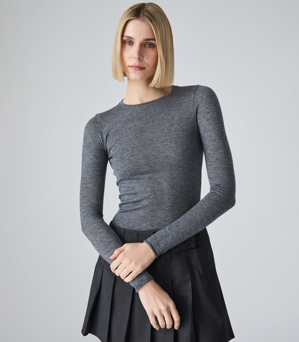 A woman with blonde bob-cut hair wearing a fitted dark gray long-sleeve top and a dark pleated skirt, standing against a light gray studio background.