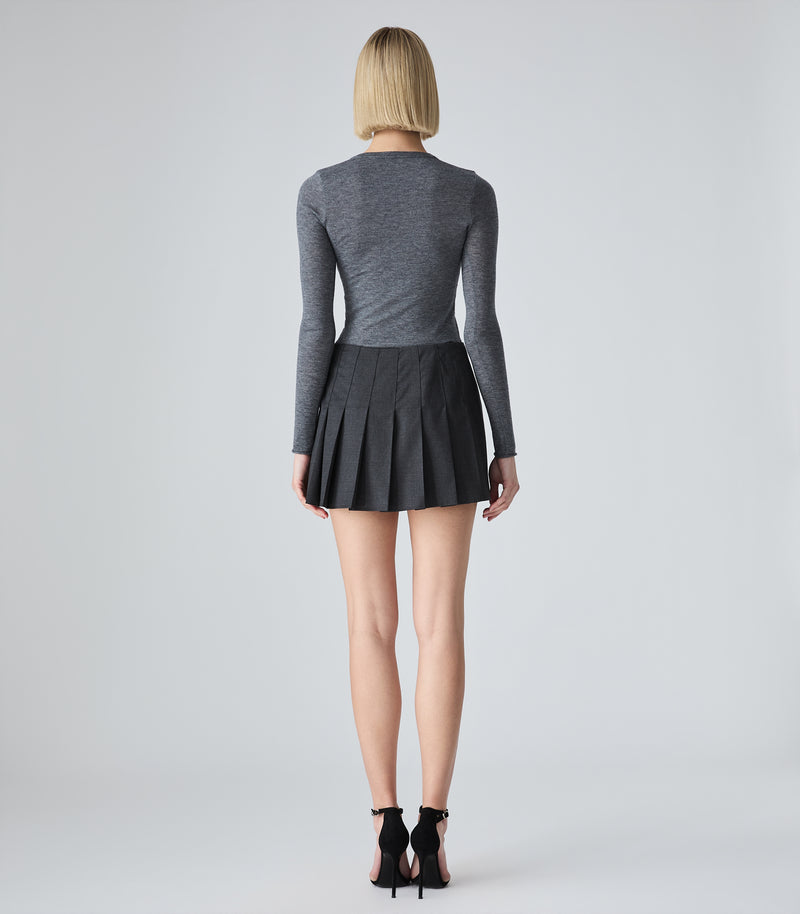 A woman viewed from behind wearing a fitted heather gray long-sleeve top and a black pleated mini skirt, paired with black high heels, against a light gray studio background.