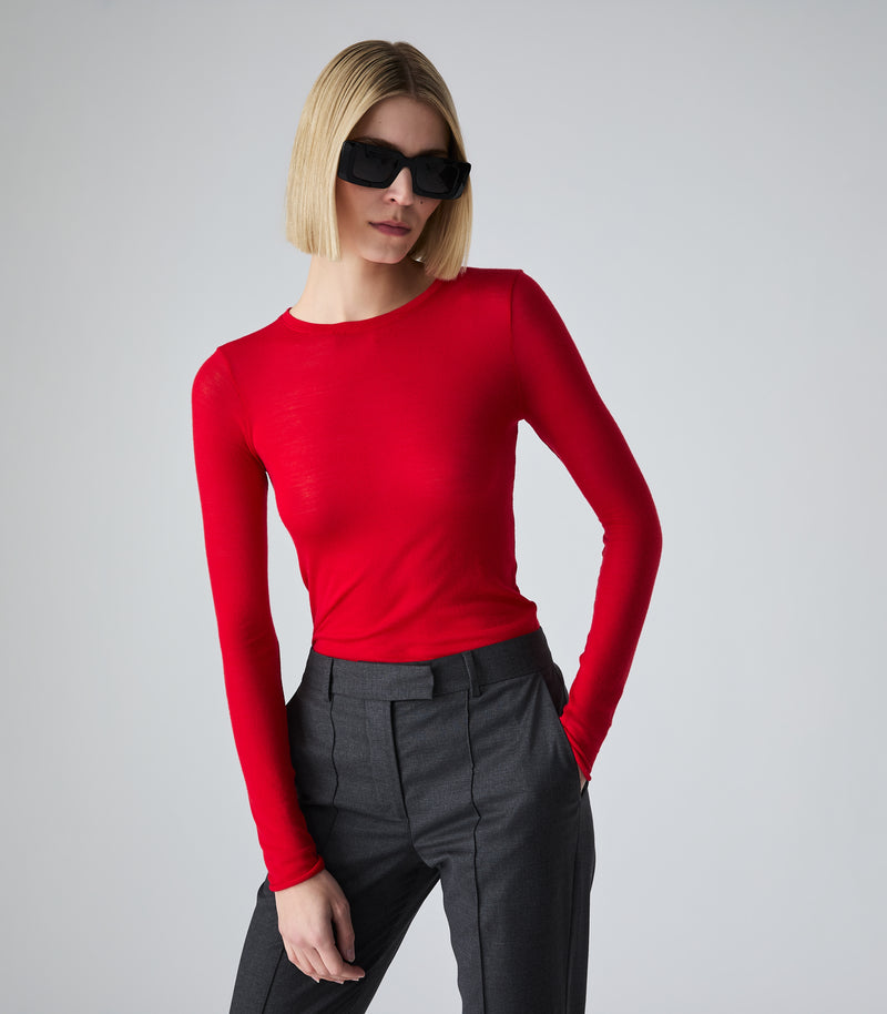 A model with a blonde bob and black sunglasses wears a bright red long-sleeve crewneck top tucked into dark gray trousers, posed against a light gray studio background.