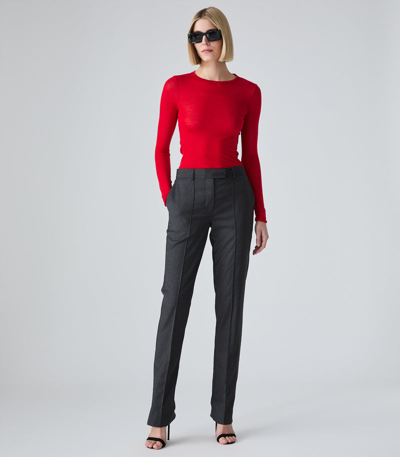 A model wearing a vibrant red long-sleeve knit top, dark charcoal gray pinstripe trousers, and black sunglasses, standing against a light gray studio background.