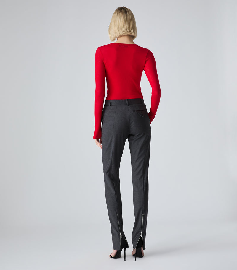 A woman viewed from the back wearing a bright red long-sleeve top and dark charcoal gray trousers with silver zipper details at the ankles, paired with black high heels, against a light gray studio background.