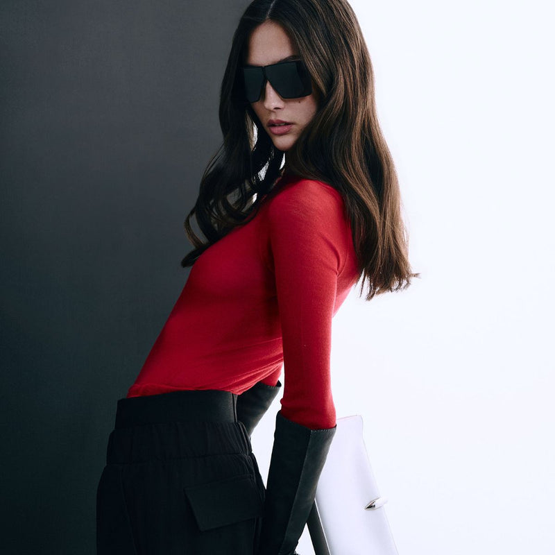 A woman wearing a fitted long-sleeve red top and black trousers, shown from the shoulders to the hips against a grey studio background.