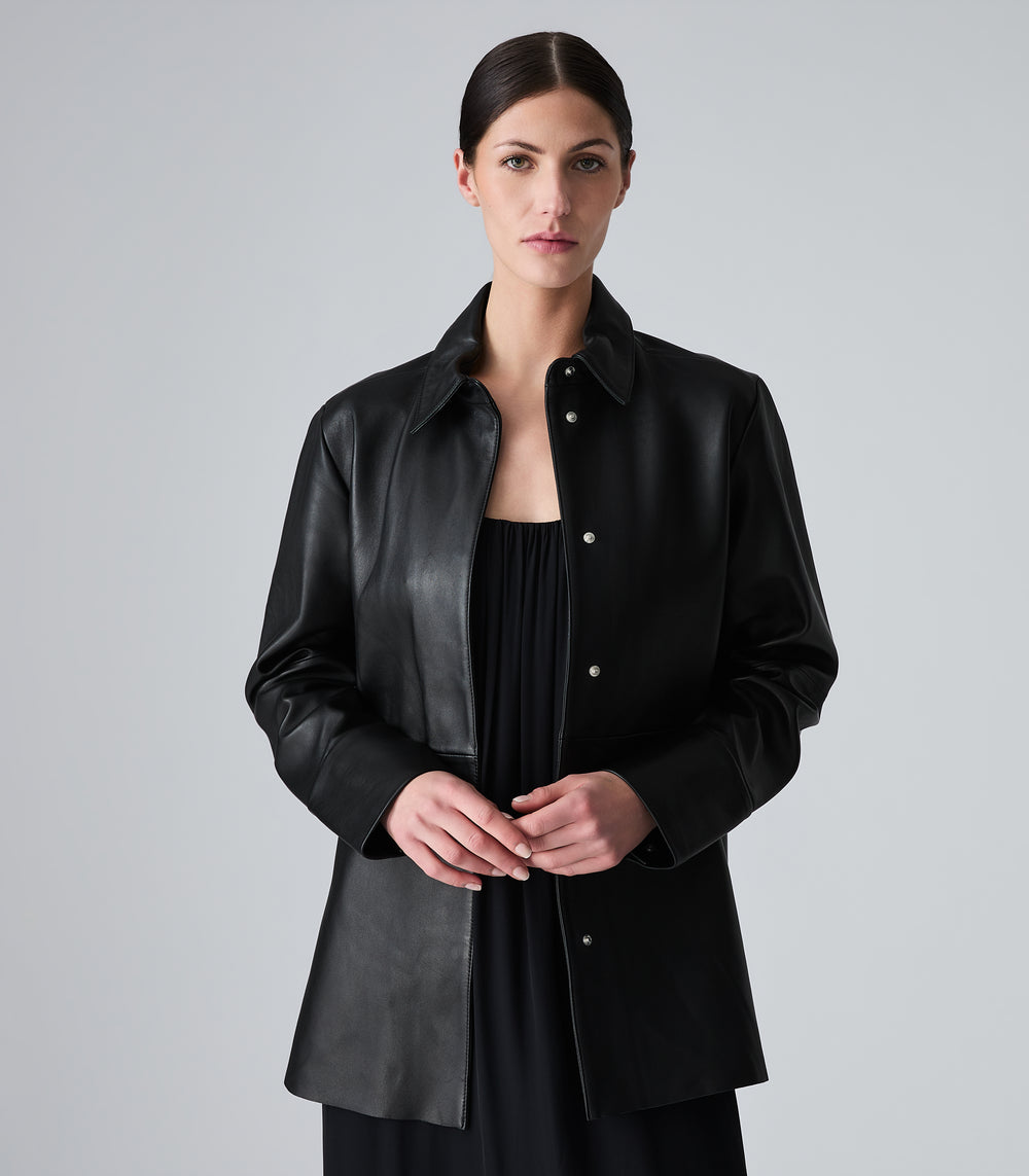 A woman wearing a black leather button-up shirt jacket over a black top, photographed against a neutral light gray studio background.