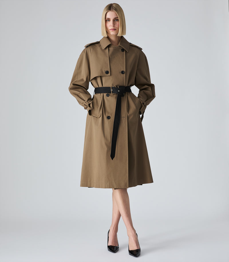 A woman modeling a classic trench coat in a warm brownish-beige color with a black belt, standing against a light gray studio background.