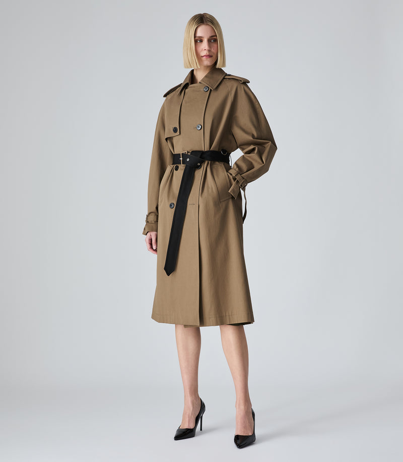 A model wearing a mid-length brown trench coat with a black belt, styled with black pointed-toe heels against a light gray studio background.