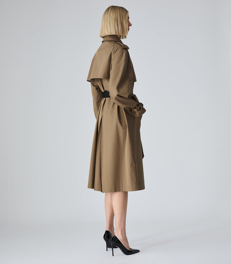 A woman modeling a mid-length brown trench coat with a matching waist belt, shown from the back in profile against a light gray studio background.