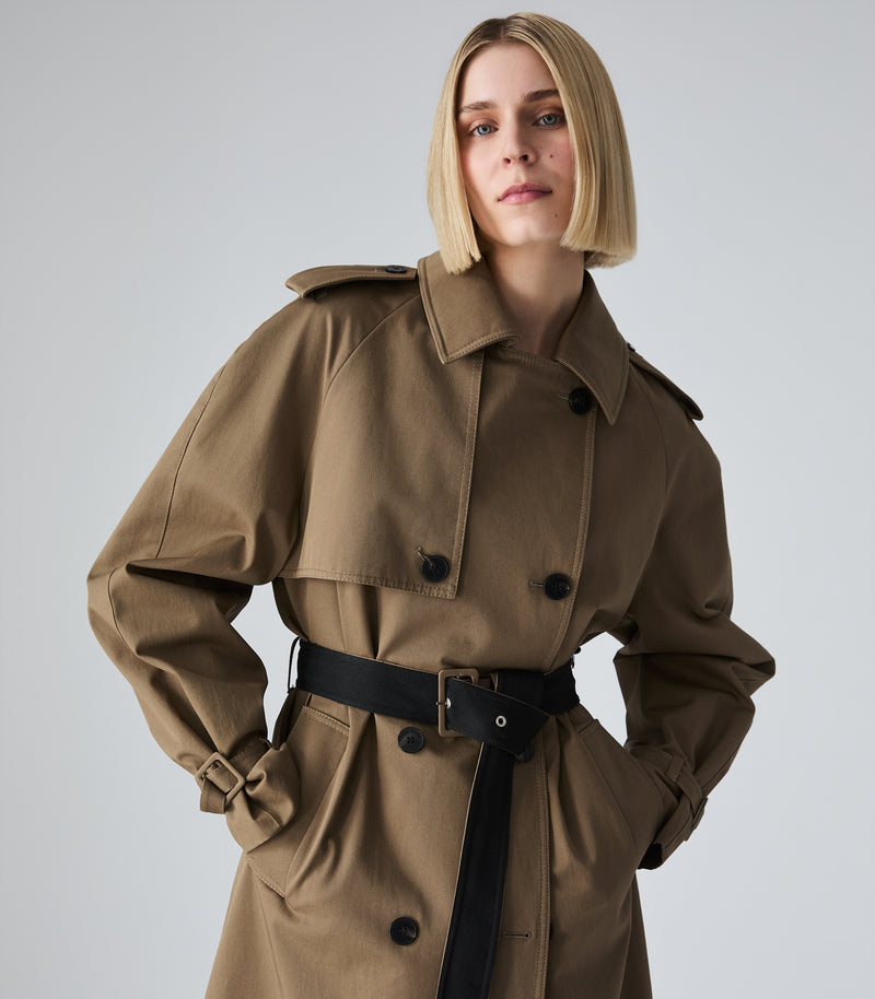 A woman wearing a classic double-breasted trench coat in brown with a black belt, photographed against a neutral light gray studio background.