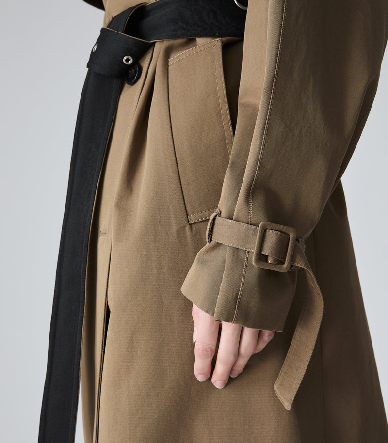 Close-up of a brown trench coat with a black belt and a hand resting near the cuff, set against a neutral gray background.