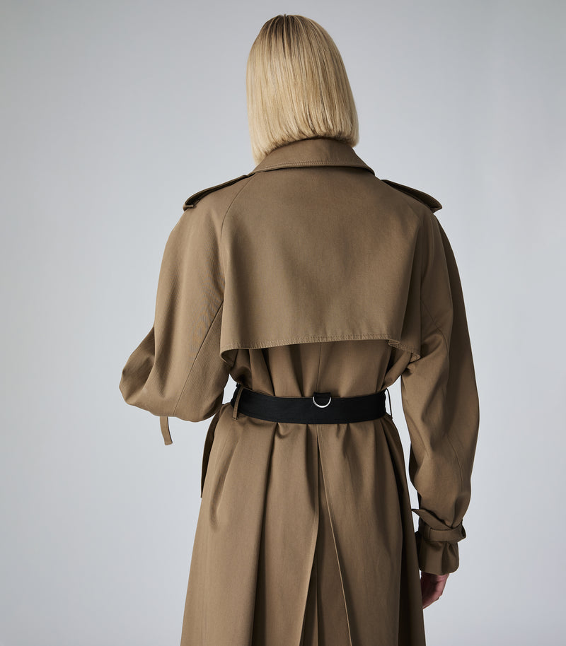 A model viewed from behind wearing a classic trench coat in camel brown with a black belt, set against a plain light gray studio background.