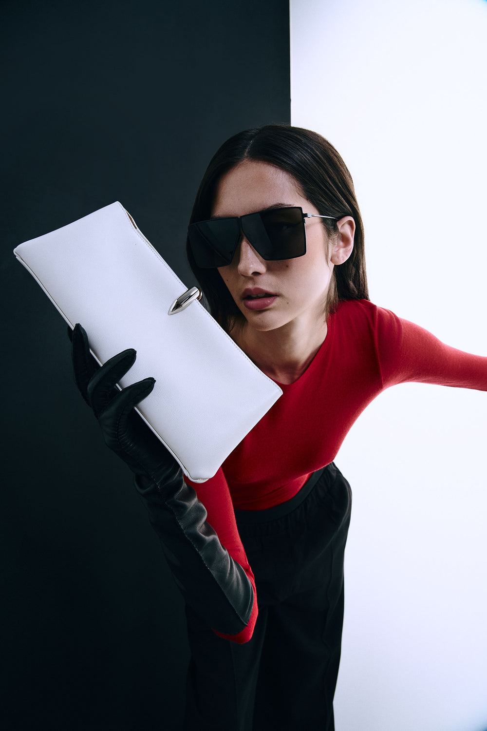 A woman in a red and black outfit, black gloves, and oversized black sunglasses holds a white rectangular clutch with a silver ring clasp against a split black and white background.