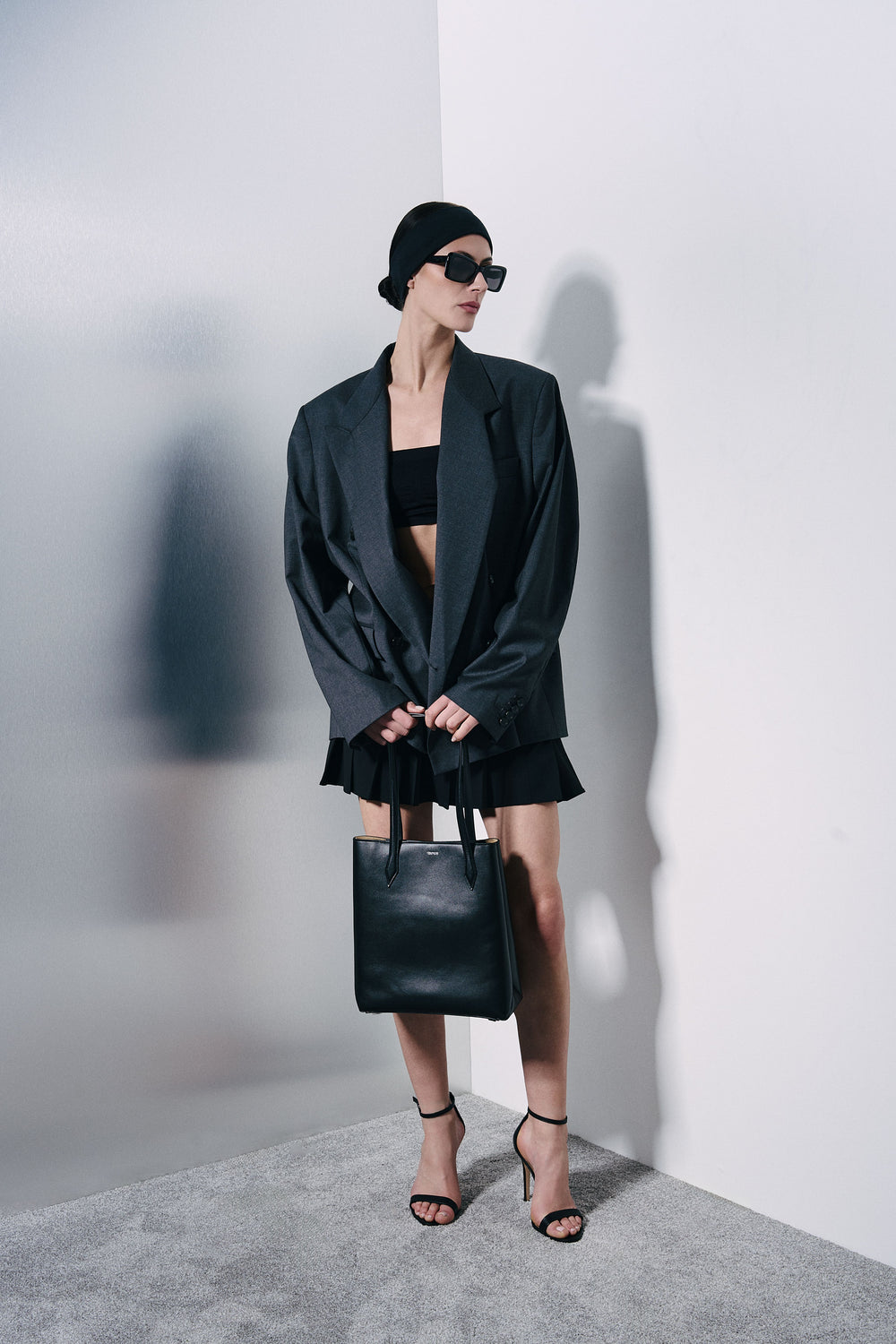A model wearing a black oversized blazer, black crop top, black pleated skirt, black beanie, black sunglasses, and black strappy heels, holding a black leather tote bag against a minimalist gray and white studio background.