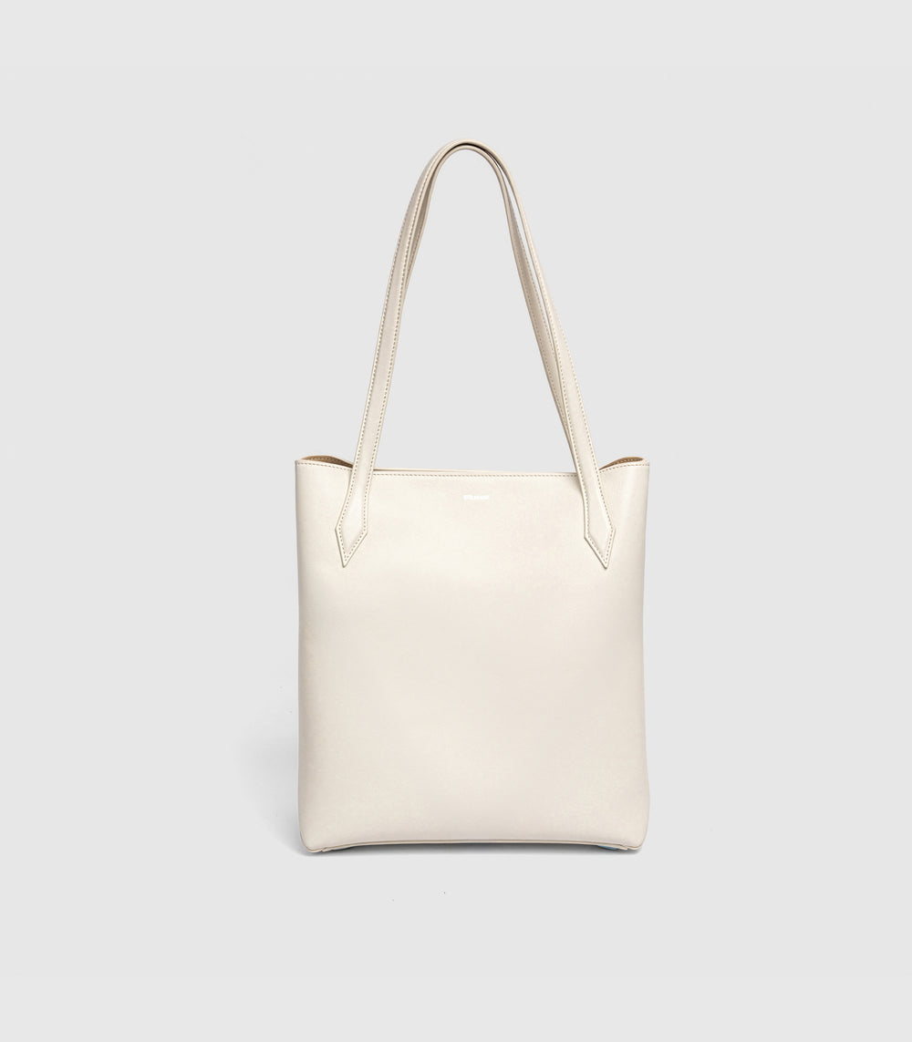 An off-white leather tote bag with long shoulder straps, shown against a light gray studio background.