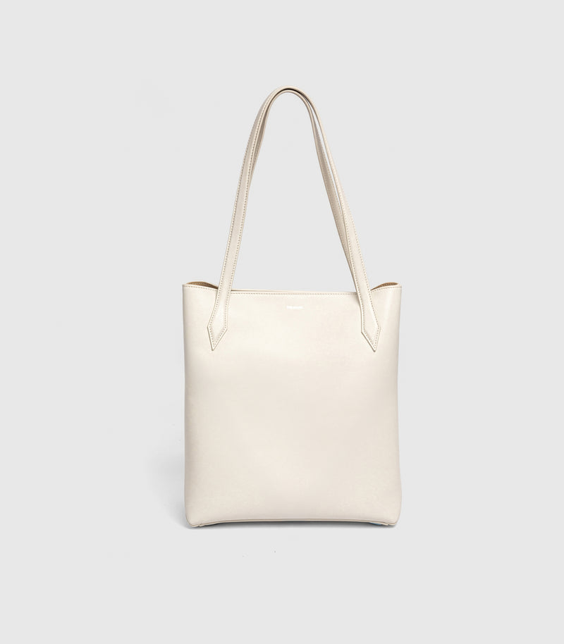An off-white leather tote bag with long shoulder straps, shown against a light gray studio background.